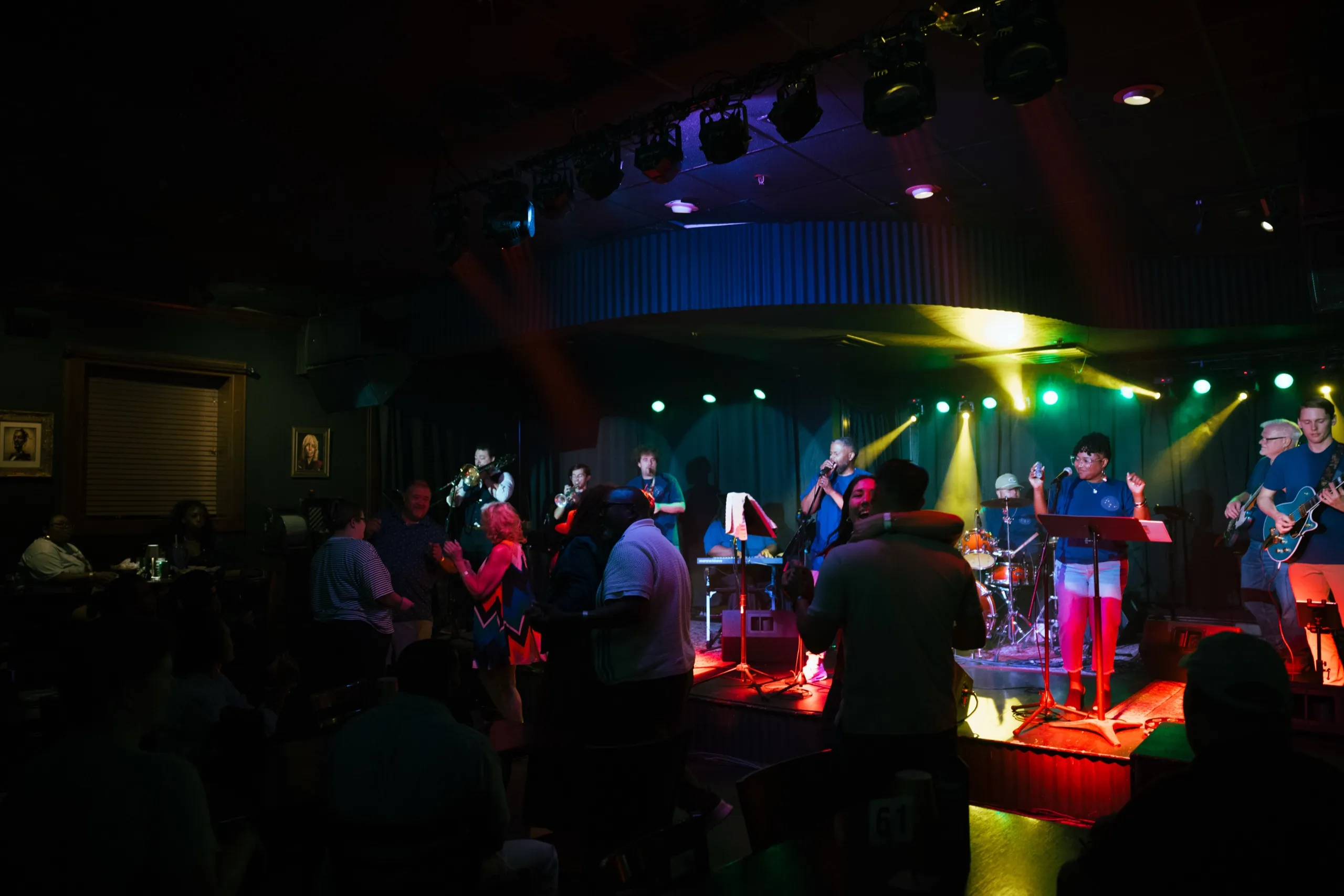 Constellation Blue performing on a red-lit club stage — horns at left, vocals centre, guitar and bass at right.